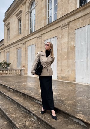 Mujer con abrigo beige y falda negra está de pie en escaleras de piedra frente a un edificio histórico con persianas blancas cerradas.