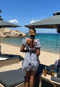 Gingham-Muster Sommerkleid mit Puffärmeln und offenem Rücken, kombiniert mit einem Strohhut, steht in der Nähe eines Strandes mit klarem Wasser und felsiger Küste.