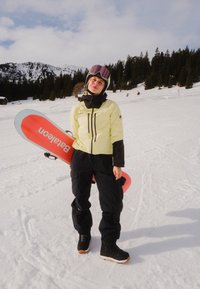 Personne portant une veste jaune et un pantalon noir tenant un snowboard rouge, debout sur une pente enneigée avec des arbres et des montagnes en arrière-plan.
