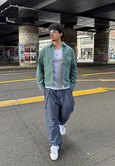 Chaqueta verde esmeralda con cremallera sobre una camiseta blanca sin mangas, combinada con pantalones grises holgados y zapatillas blancas. Incluye gafas de sol con tintado azul y un detalle de cadena.