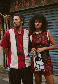 Camiseta a rayas rojas y blancas con una cámara, pantalones negros; mujer con un vestido rojo de corazones estampados con mangas con volantes, sosteniendo un bolso en blanco y negro.