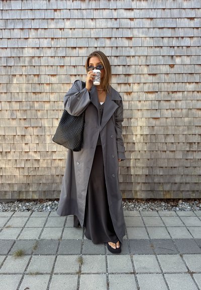 Abrigo gris de trench con grandes botones, llevado sobre una blusa gris y una falda larga. Accesorios incluyen un bolso de tela negro y gafas de sol. Sosteniendo una taza de café.