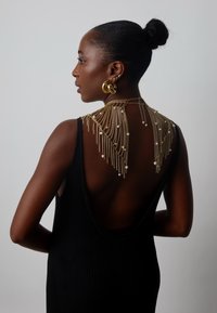 Czarna prążkowana sukienka z odkrytymi plecami, ozdobiona złotymi łańcuszkami i perłami opadającymi na ramiona. Look dopełniają okrągłe złote kolczyki.