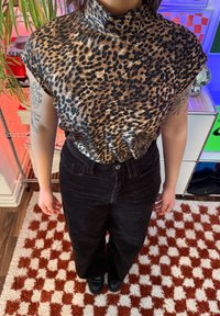 Personne portant un haut imprimé léopard et un pantalon noir, debout sur un tapis à carreaux rouge et blanc dans une pièce colorée.