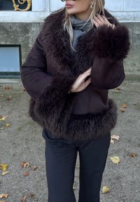 Smeđa jakna od umjetnog krzna s mekanim ovratnikom i manšetama, s glatkim tkanim dijelom tijela i pripijeni krojem. Uparena s crnim hlačama.