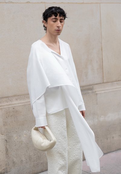 Persona con cabello rizado y oscuro que lleva una camisa blanca extragrande con tela larga y drapeada, pantalones blancos texturizados, sosteniendo un pequeño bolso crema.