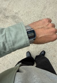 Osoba v zelenej bunde a sivej košeli kontroluje strieborné digitálne hodinky Casio, zatiaľ čo stojí na betóne. Vidieť čierne nohavice a topánky.