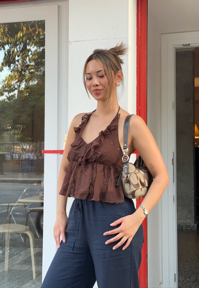 Jeune femme avec un chignon portant un haut à volants marron à col licou et un pantalon bleu marine, debout dehors près d'une porte en verre et d'une fenêtre, portant un sac à main à motifs.