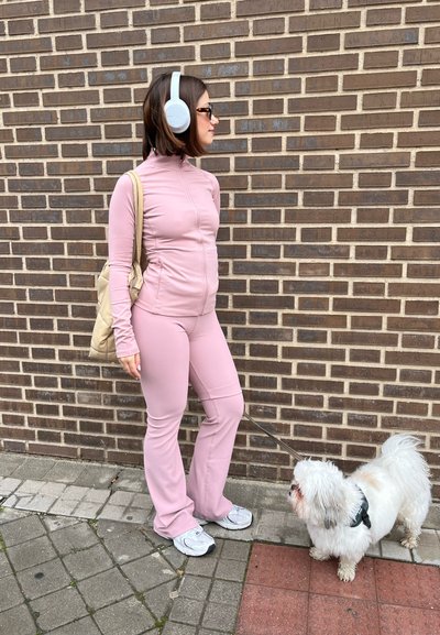 Chaqueta rosa de cremallera y pantalones a juego con textura acanalada; zapatillas blancas; llevando un bolso de color beige claro; de pie junto a un pequeño perro blanco.