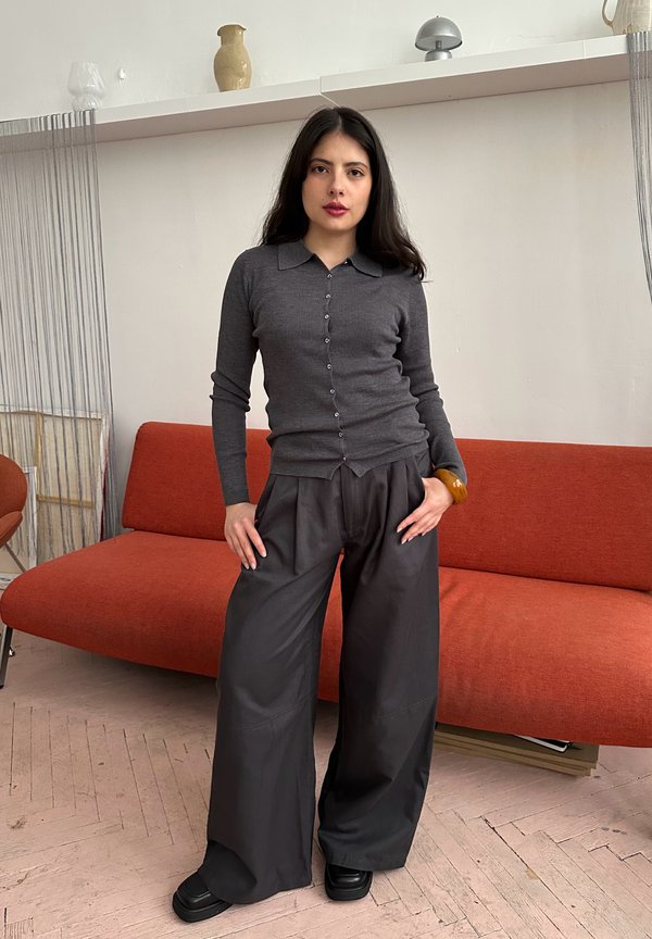 Mladá žena s dlhými tmavými vlasmi, oblečená v sivom kardigane na gombíky a širokých čiernych nohaviciach, stojaca pred oranžovou pohovkou.