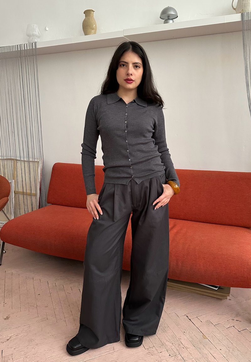 Jeune femme aux longs cheveux foncés portant un gilet gris boutonné et un large pantalon noir, debout devant un canapé orange.