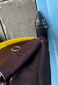 Μαύρες μυτερές μπαλαρίνες με διπλές ασημένες πόρπες, συνδυασμένες με μια καφέ υφασμάτινη τσάντα με κίτρινη λωρίδα, τοποθετημένη πάνω από σκούρους παντελόνες.