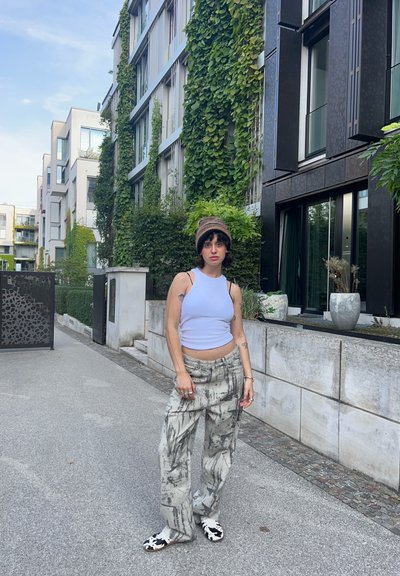 Una camiseta sin mangas ajustada de color azul claro, pantalones holgados grises con estampado y zapatos tipo slip-on en blanco y negro. Follaje verde y edificios modernos de fondo.