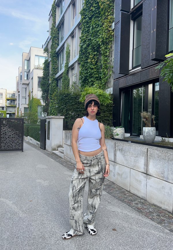 Een lichtblauw aansluitend tanktop, grijze baggy broeken met een patroon en zwart-witte instappers. Groene loofbomen en moderne gebouwen op de achtergrond.