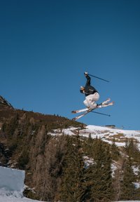 Un skieur portant une veste noire et un pantalon blanc saute en l'air avec des skis multicolores, avec de la neige et des arbres visibles en arrière-plan sous un ciel bleu clair.