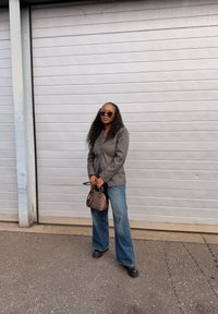 Γυναίκα με μακριά, σγουρά μαλλιά που φορά γυαλιά ηλίου, γκρι σακάκι, τζιν παντελόνι φαρδιάς γραμμής, μαύρα παπούτσια, κρατώντας μια μικρή καρό τσάντα, στέκεται έξω.