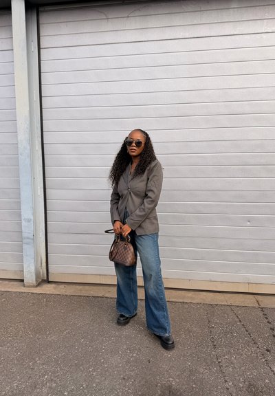 Mujer con cabello largo y rizado, lleva gafas de sol, blazer gris, vaqueros de pierna ancha, zapatos negros, sostiene un pequeño bolso de cuadros, de pie al aire libre.