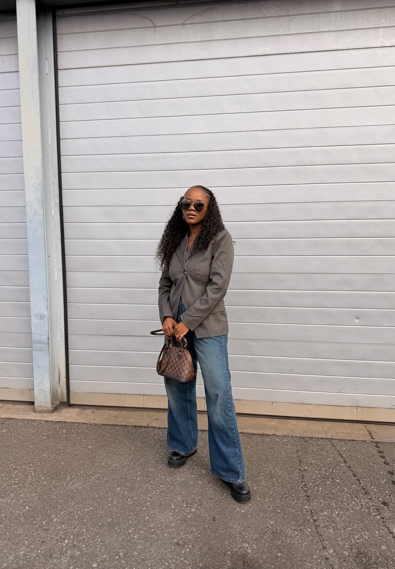 Kvinde med langt krøllet hår iført solbriller, grå blazer, vidde jeans, sorte sko, holder en lille ternet håndtaske, står udendørs.