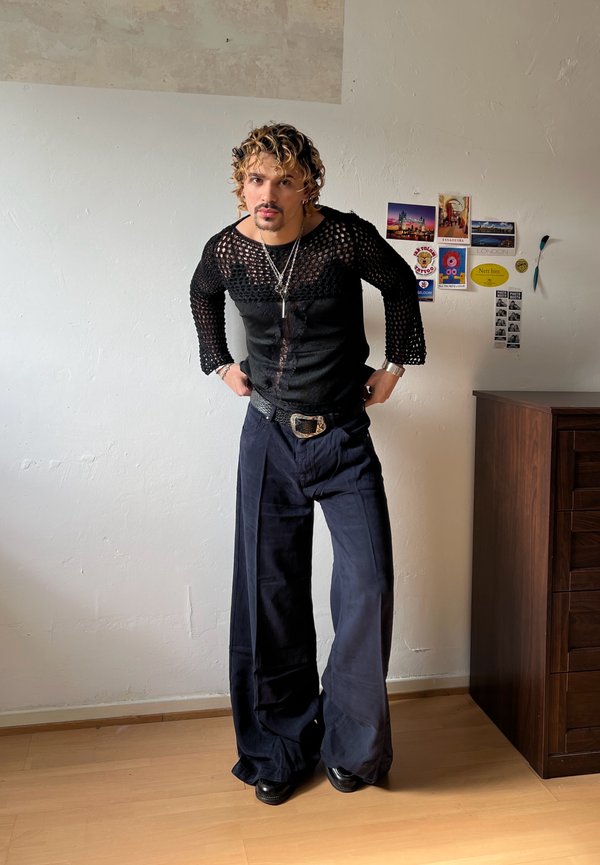 Bărbat cu păr creț, purtând un top negru din plasă cu mâneci lungi și pantaloni bleumarin largi, stă înăuntru aproape de un perete cu cărți poștale și o comodă din lemn.