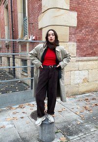 Μπεζ παλτό σεχάρτη πάνω από κόκκινη μπλούζα, σκούρες καφέ μπορντούρες παντελόνια και γκρι αθλητικά παπούτσια. Στέκεται πάνω σε τσιμεντένιο βάθρο μπροστά σε τοίχο από τούβλα.