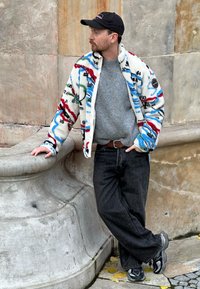 Veste en polaire floue, multicolore et à motifs avec des accents noir et bleu, pull gris, jeans noirs et baskets grises. Fond en pierre neutre.