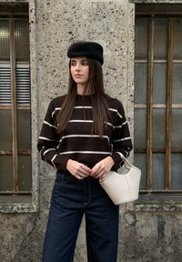 Femme aux longs cheveux bruns portant un béret noir, un pull rayé et un jean foncé, tenant un petit sac à main blanc contre un mur en pierre.