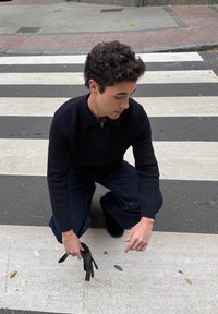 Homme agenouillé sur un passage pour piétons, tendant la main vers le sol et tenant un gant noir dans une main, portant un pull sombre et un jean.