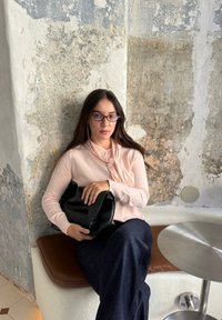 Sac noir tenu à la main, haut à manches longues rose pâle avec détail noué, jean bleu foncé à jambes larges, assis sur un coussin marron contre un mur texturé.