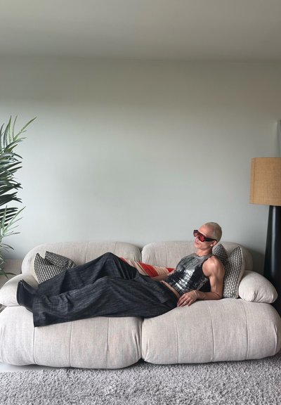 Persona con cabello rubio corto y gafas de sol rojas descansando en un sofá beige, vistiendo un top negro de lentejuelas y pantalones de pierna ancha en una sala de estar minimalista.