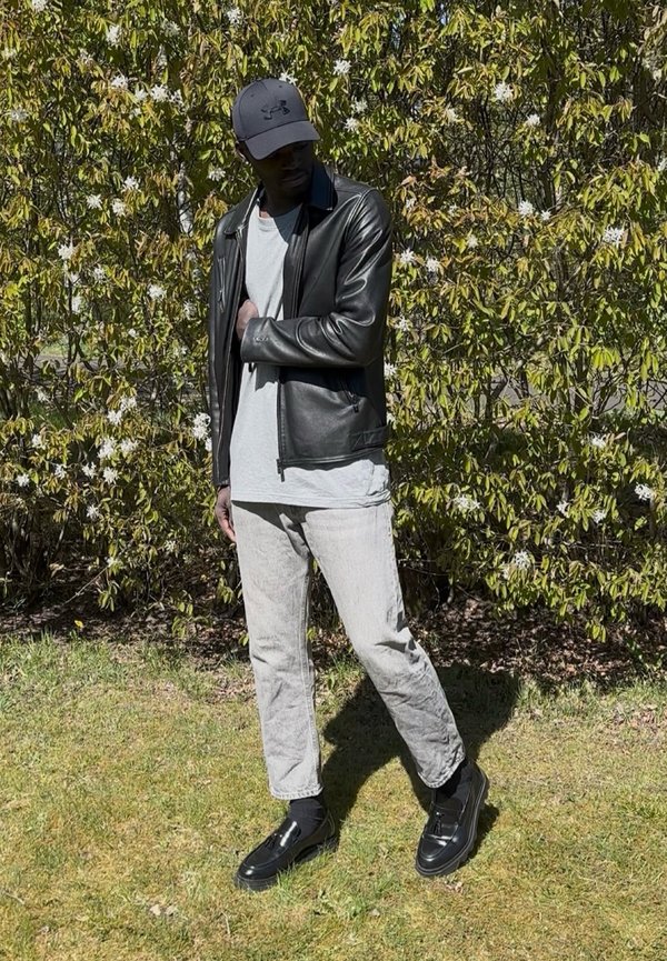 Mann trägt schwarze Lederjacke, hellgraue Hose, schwarze Schuhe und dunkle Kappe, steht auf Gras vor einem grünen Busch mit weißen Blumen.