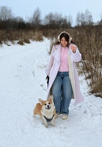 Femme en manteau blanc et cache-oreilles promenant un corgi souriant sur un chemin enneigé avec de l'herbe sèche et des arbres dépouillés à proximité.