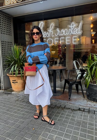 Top de rayas azul y marrón de manga larga, falda asimétrica blanca, bolsa de mimbre con acentos en rojo y marrón claro, y sandalias negras con correas decorativas.