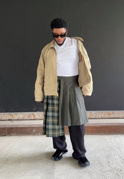 Chaqueta beige sobre una camisa blanca, combinada con una falda de cuadros verde y a rayas. Pantalones sueltos negros y zapatos completan el conjunto.