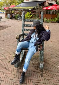 Grote groene metalen schommelstoel in een park, persoon zittend in een spijkerjack, blauwe jeans, zwarte laarzen, met een bloemenhandtas in de hand. Pad zichtbaar.