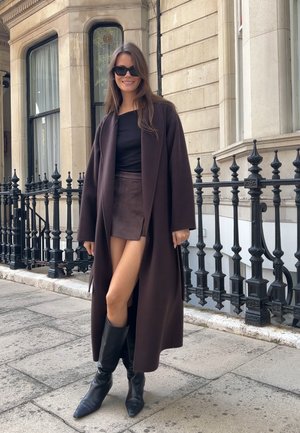 Abrigo largo marrón sobre una blusa negra y una falda corta marrón. Botas negras hasta la rodilla. Calle adoquinada y barandilla de hierro forjado de fondo.