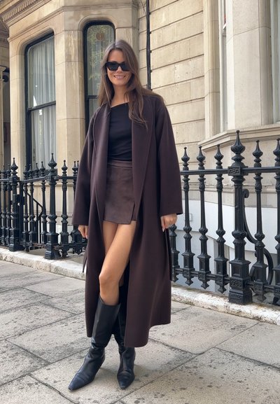 Abrigo largo marrón sobre una blusa negra y una falda corta marrón. Botas negras hasta la rodilla. Calle adoquinada y barandilla de hierro forjado de fondo.