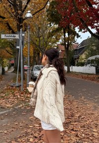 Jersey grueso de punto color crema con un patrón de trenzas, acompañado de una bufanda de cuadros beige, usado sobre pantalones cortos blancos en una calle cubierta de hojas.