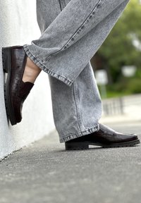 Σκούρες καφέ loafers με ανάγλυφο σχέδιο κροκοδείλου και χαμηλό τακούνι, φορεμένα με ανοιχτό γκρι παντελόνι φαρδιάς γραμμής, που αναδεικνύουν μια επίπεδη καουτσούκ σόλα και λεία επιφάνεια.