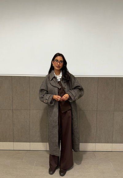 Mujer con cabello largo y oscuro y gafas, que lleva un abrigo largo de cuadros sobre pantalones marrones y chaqueta, de pie contra una pared de azulejos bicolor.