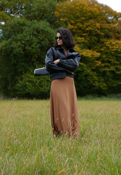 Chaqueta bomber de cuero negra, vestido maxi beige y bolso negro; de pie en hierba alta con follaje verde y amarillo al fondo.
