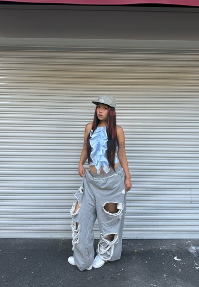 Top azul claro con volantes, sudaderas gris desgastadas con grandes roturas, zapatillas blancas y una gorra gris clara con logo, contra un fondo texturizado.