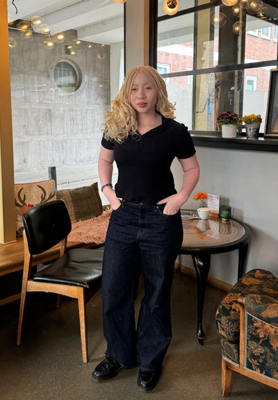 Camisa polo negra de canalé, jeans de pierna ancha azul oscuro y zapatos negros. El fondo presenta una ventana redonda, cojines decorativos y una mesa con plantas.