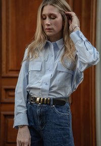 Lyseblå skjorte med knapper og to brystlommer, kombinert med mørkeblå denimjeans og et svart belte med gulldetaljer.