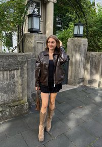 Giacca di pelle marrone, top nero in cotone, gonna di pizzo, stivali da cowboy color beige, mentre tiene una borsa marrone; in piedi su un pavimento di pietra in un ambiente verde.