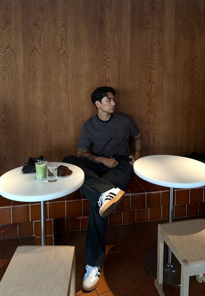 Camiseta a rayas, pantalones anchos negros y zapatillas blancas con rayas negras. Sentado en una mesa blanca con bebidas y un pastelito. Fondo de panel de madera.