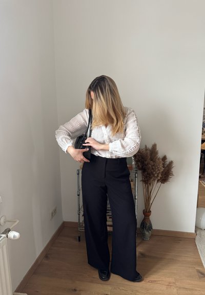 Mujer con cabello rubio que lleva una blusa blanca y pantalones negros de pierna ancha, ajustando la correa de un bolso negro en una habitación minimalista.