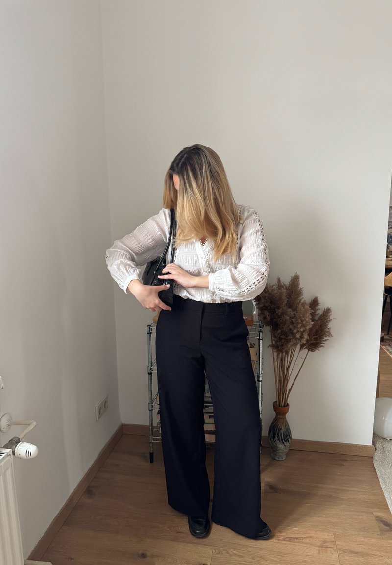 Femme aux cheveux blonds portant un chemisier blanc et un pantalon large noir, ajustant la lanière d'un sac à main noir dans une pièce minimaliste.