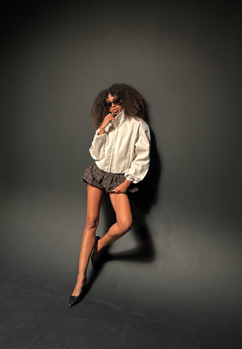 Femme aux cheveux bouclés portant des lunettes de soleil, une veste blanche, une jupe courte à carreaux et des talons noirs, posant sur un fond sombre.