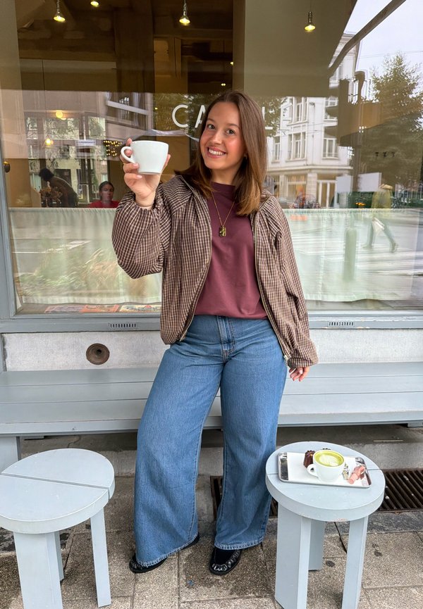 Joven mujer sonriendo al aire libre sosteniendo una taza blanca con café, parada junto a dos mesas pequeñas, en una hay una taza y postres.
