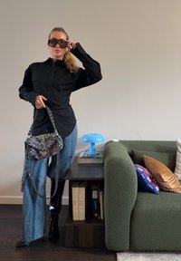Femme en chemise noire, jean bleu et bottes noires tenant un sac à main coloré, debout à côté d'une étagère sombre et d'un canapé vert avec des coussins.